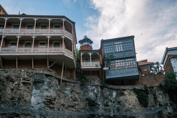 Traditional Georgian houses with colorful carved wooden balconies perched on the Metekhi cliff edge above the Kura river in the old town of Tbilisi, Georgia. Historic architecture at sunset. © Michele