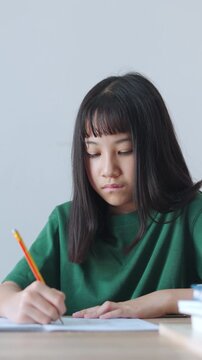 Vertical video close up and selective focus showing Asian kid girl concentrating as she writes in her notebook with a pencil. Bright daylight fills the room while she is doing her homework.