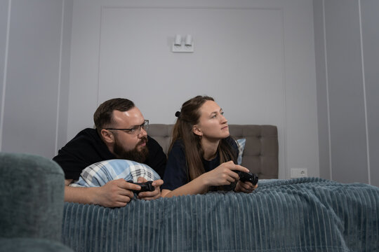 Young heterosexual couple lying comfortably in bed, playing video games with wireless controllers, experiencing leisure, entertainment, and relaxation in a cozy home environment