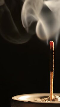 a single lit incense stick. Thin white smoke rises slowly and curls from the glowing tip against a very dark background. Minimal warm lighting. No hands.