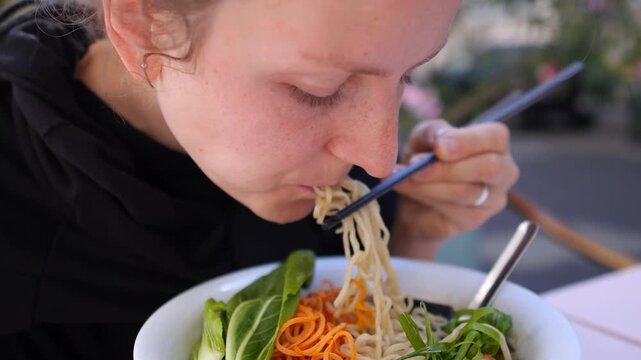 Close up of a young woman enjoying a healthy bowl of traditional asian ramen soup, using chopsticks to eat the fresh noodles with vegetables and tofu at an outdoor restaurant