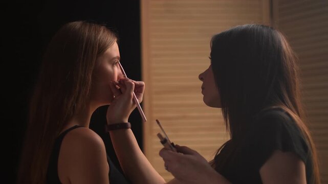 Professional makeup artist carefully applying concealer under a beautiful model's eye with a brush in a beauty salon, highlighting the skin for a photoshoot session