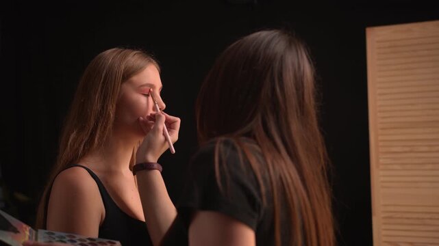 Professional makeup artist carefully applying pink eyeshadow with a brush to a young female model's eyelid in a beauty salon, creating a vibrant look for a fashion event