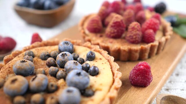 Two fresh homemade fruit tartlets with custard, one decorated with raspberries and the other with blueberries and cinnamon powder, shifting focus from one to the other on a wooden board