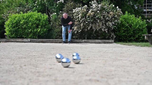 Man playing petanque on sunny park court with boules scattered on gravel, casual outdoor leisure and recreation game in European setting