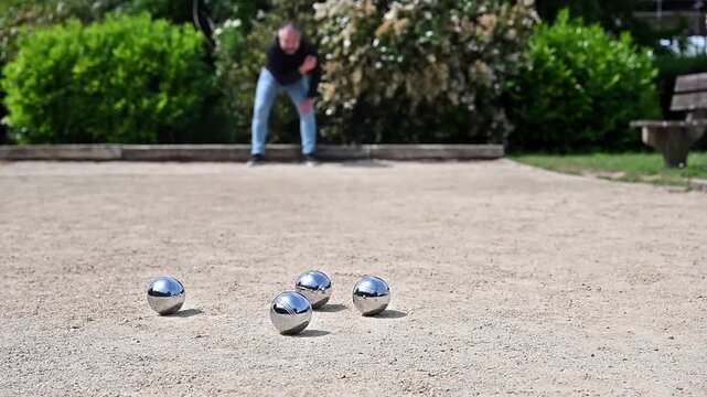 Man throwing petanque ball on gravel court wide view with green park surroundings, full body outdoor leisure sport action and game scene