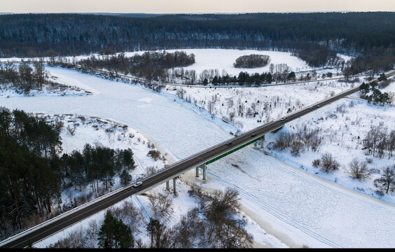 Aerial view of a bridge crossing a frozen river surrounded by snow-covered fields and dense pine forests in Lithuania.