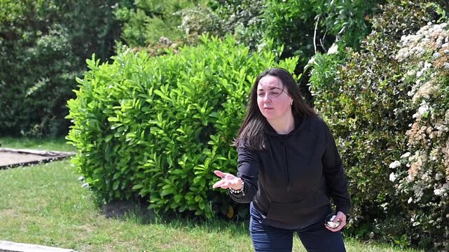 Woman playing petanque in slow motion on gravel court holding spare ball in other hand, focused outdoor leisure and recreation moment