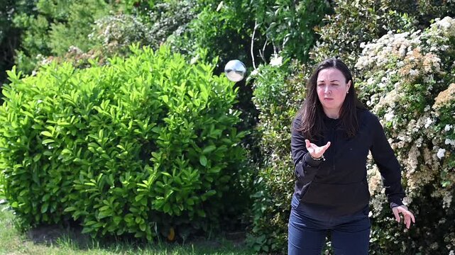 Woman throwing steel petanque ball in slow motion on sunny park court, graceful underhand release and ball trajectory in outdoor leisure game