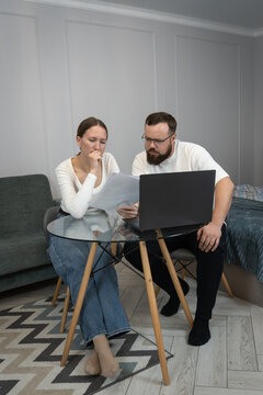 Young couple discussing financial documents, managing personal budget, and making online payments using a laptop in their home living space, focusing on budgeting and debt awareness. Vertical photo
