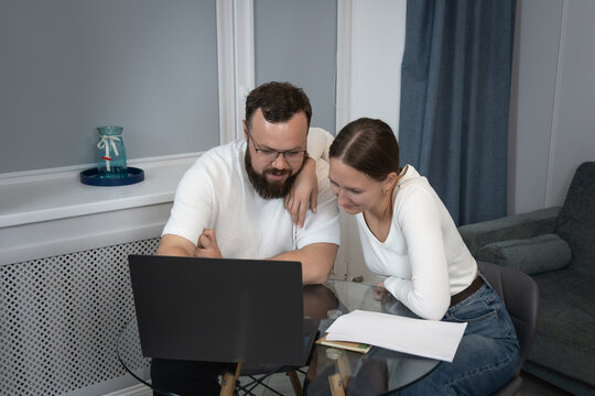 Couple collaborating while looking at a laptop screen, discussing information and planning finances or remote work from their apartment, representing teamwork and modern lifestyle