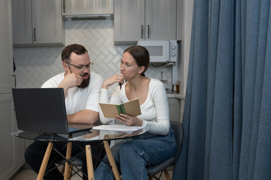 Young couple planning household finances at their kitchen table, using laptop and notebook to budget bills, savings and future expenses while discussing strategy together