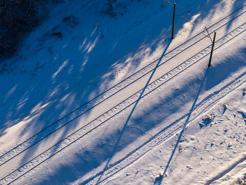 Aerial view of railway tracks covered in fresh snow with long tree shadows and power line poles during winter in Lithuania.