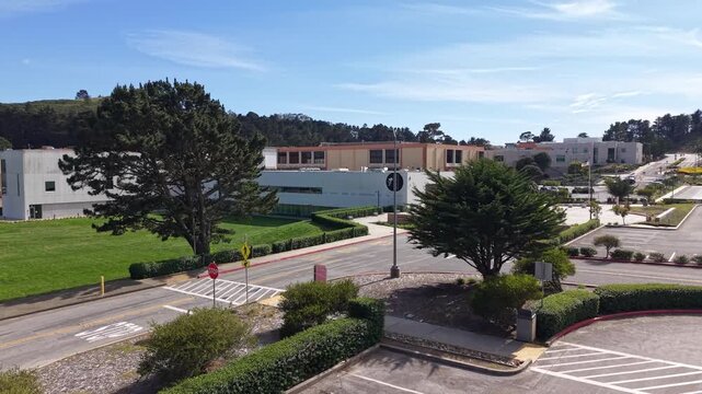 Ascending drone view over the empty parking lot and modern buildings of Skyline College campus in San Bruno, California, on a bright sunny afternoon with blue sky and green landscape