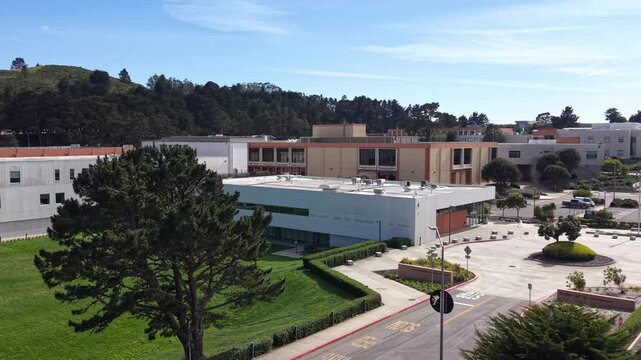 Aerial drone circling Skyline College campus in San Bruno, California, showcasing modern architecture, green lawns, parking areas and surrounding hills under a clear blue sky