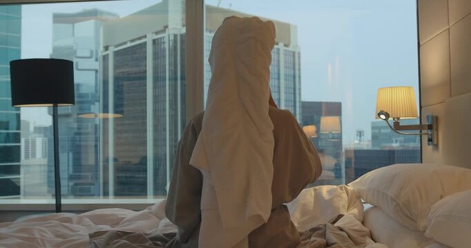 Woman wearing bathrobe and head towel sitting on bed drinking coffee while looking at city skyscrapers through hotel window. Morning routine in luxury urban apartment from back view.