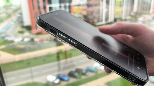 Hand holds a smartphone near a window. The phone tilts and the thumb taps the touchscreen over a blurred city view. Mobile app browsing and digital interaction concept..