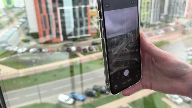Hand holds smartphone by a window above the city. The phone records an urban view in close up with a mobile camera interface. Content creation and travel photography concept..