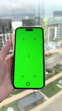 Hand holds smartphone with green screen near city window. Thumb taps on a mobile mockup in front of blurred urban buildings. Advertising template for app design and digital content..