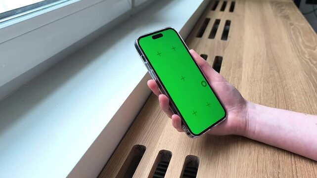 Hand holds smartphone with green screen beside window. Close up mobile device on wooden desk with copy space. App mockup, advertising, and editing concept..