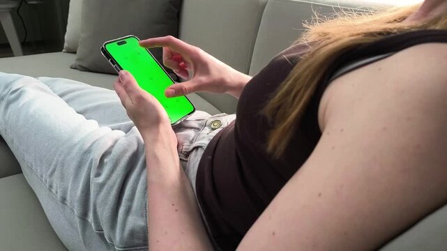 Woman lies on sofa and zooms image on smartphone using two fingers at home. Female performs pinch gesture on mobile screen. Concept image scaling, touch interaction, digital usage, modern