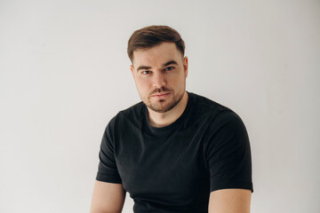 Obraz premium Male model with short brown hair wearing a black t-shirt poses against a plain white background, showcasing a confident expression and relaxed posture