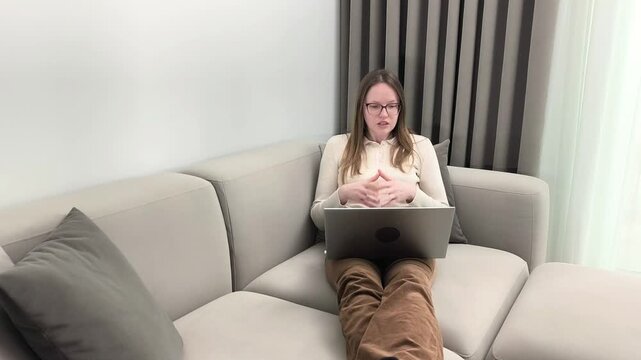 Woman sits on sofa with laptop and gestures explaining business project. Female communicates ideas through online conversation using expressive hand movement. Concept of remote presentation