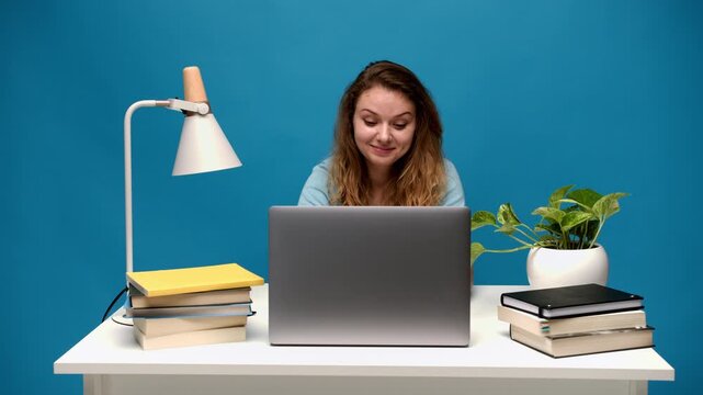 Young student smiling and typing on a laptop computer while studying online with books against a blue background