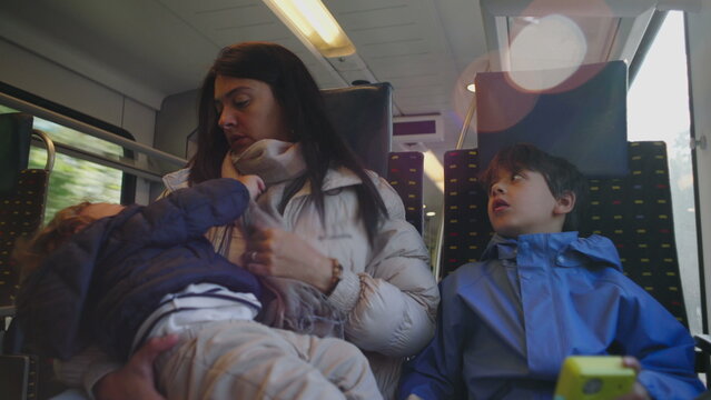 Mother sits on train holding restless toddler while older child looks upward creating authentic moment of overwhelmed parenting family travel fatigue and real everyday commute inside public transport