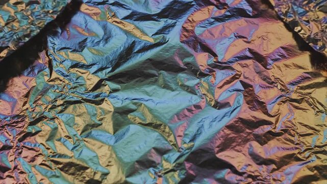 Abstract textured background of crumpled iridescent foil with shifting rainbow colors of blue, green, purple, and gold creating a vibrant and dynamic surface