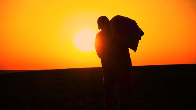Silhouette of a male farmer with a sack of wheat on his shoulder. A male worker at sunset with a sack of fertilizer in his hand. The concept of farming, agriculture, hard work of workers.