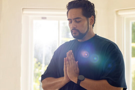Mid adult African man praying at home near windows with lens flare wearing dark tee, smartwatch