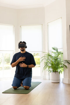 African American man kneeling on yoga mat at home, using VR headset and headphones