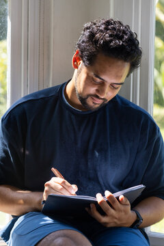 Mid-adult African American man writing in notebook at home on window seat, navy shirt, wristwatch