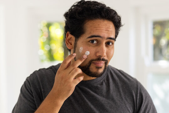 African American man applying facial cream on cheek at home, cream on fingers, gray T shirt