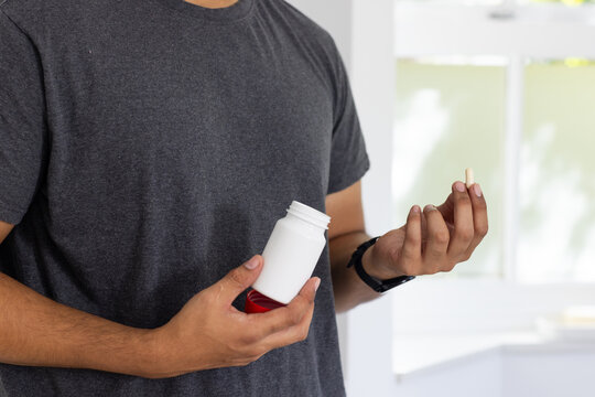 Mid adult African American man holding white pill bottle and examining beige capsule at counter