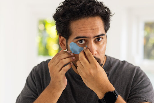 Mid-adult man applying blue crescent hydrogel under-eye patch at home vanity wearing gray T-shirt