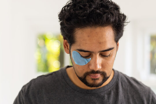 Man wearing gray t-shirt using blue under-eye gel patch beneath right eye near windows