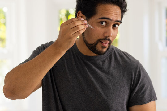 African American man wearing dark gray tee holding glass dropper applying serum to cheek at vanity