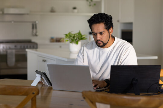 Mid-adult man working on silver laptop at wooden table in home kitchen with monitor and tablet