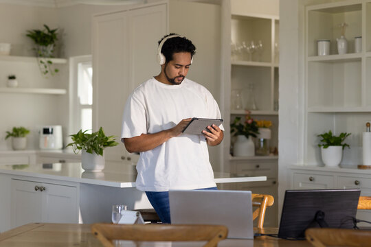 Man standing at kitchen island wearing white headphones and T-shirt, using tablet near two laptops