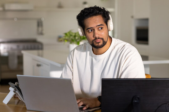 African American man using multiple devices at kitchen table wearing light crewneck and headphones