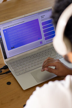 Mid-30s African man working on silver laptop at wooden table wearing white headphones, copy space