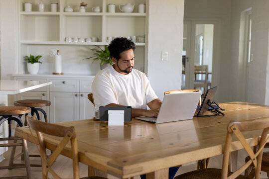 Mid adult African American man working on silver laptop at wooden dining table in kitchen