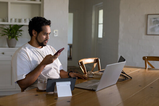 African American man multitasking on wooden dining table in kitchen, using laptop smartphone tablet