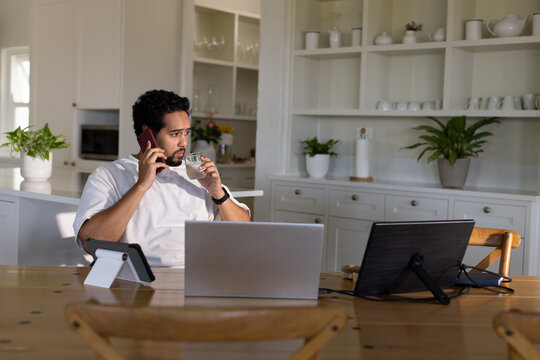 African American mid-adult man holding smartphone and glass while working on laptop in home kitchen