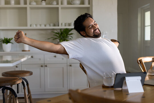 African American man in his 30s stretching back in kitchen at wooden table with tablet