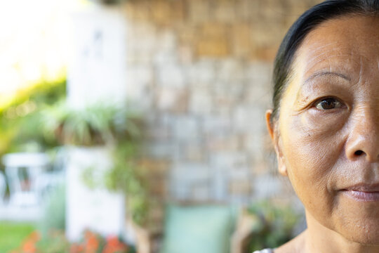 Senior Asian woman looking toward camera on patio with potted plants, patterned top, copy space