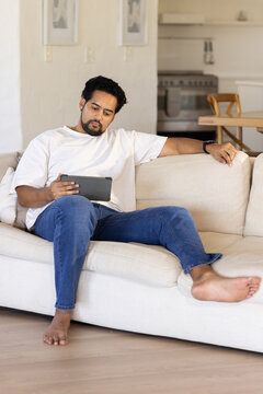 African American mid-adult man sitting barefoot on beige sofa in home living area, holding tablet