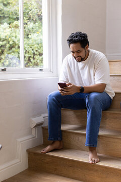 African American man sitting on wood stairs by window, in white T, jeans, watch, holding smartphone